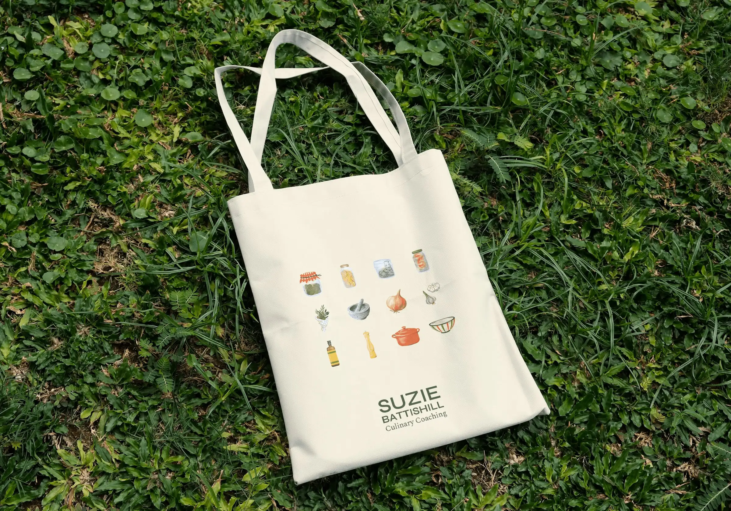 Canvas tote bag on grass, printed with Suzie Battishill’s illustrated kitchen staples and wordmark