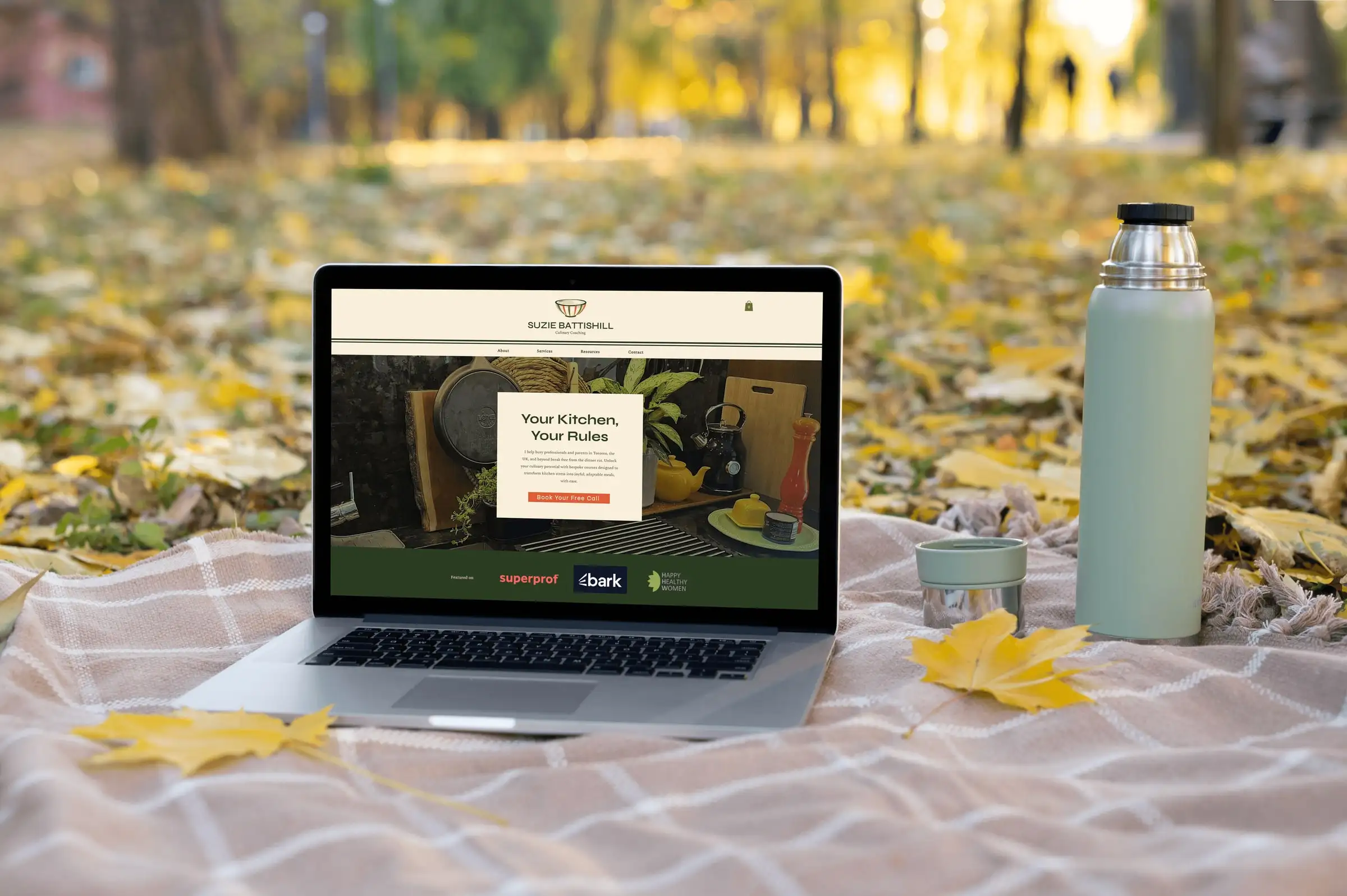 A laptop on a picnic blanket showing the Suzie Battishill homepage, headed “Your Kitchen, Your Rules”