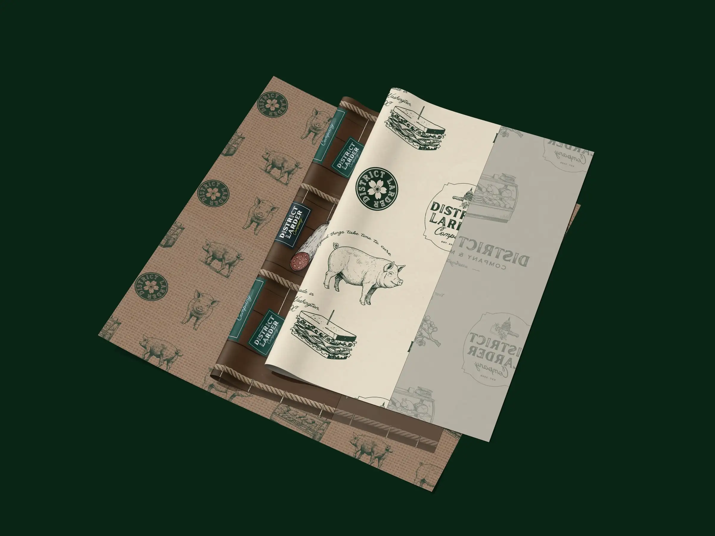 Four District Larder pattern papers fanned out: kraft with pigs and seals, dark brown, cream with pigs and sandwiches, and grey with the wordmark